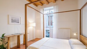 Een slaapkamer met een hemelbed, een nachtkastje met een lamp, een ingelijst schilderij aan de muur, en uitzicht op het balkon.