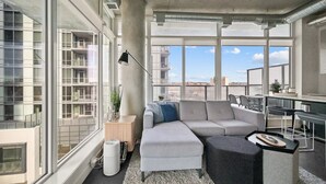 Una sala de estar moderna con un sofá seccional, una pequeña mesita auxiliar, una lámpara de pie y vistas de la ciudad a través de amplios ventanales.