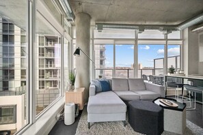 Ein modernes Wohnzimmer mit einem Ecksofa, einem kleinen Beistelltisch, einer Stehlampe und Blick auf die Stadt durch große Fenster.