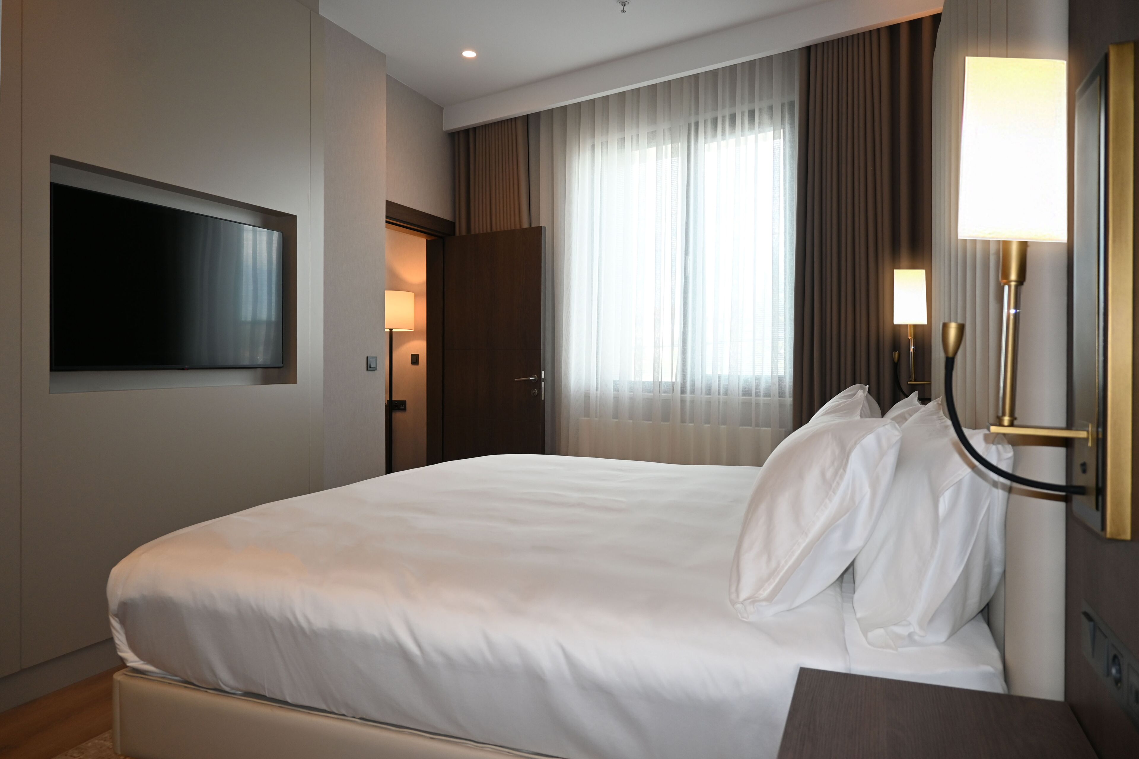 Una habitación de hotel con cama, televisor montado en la pared, mesita de noche y ventana con cortinas.