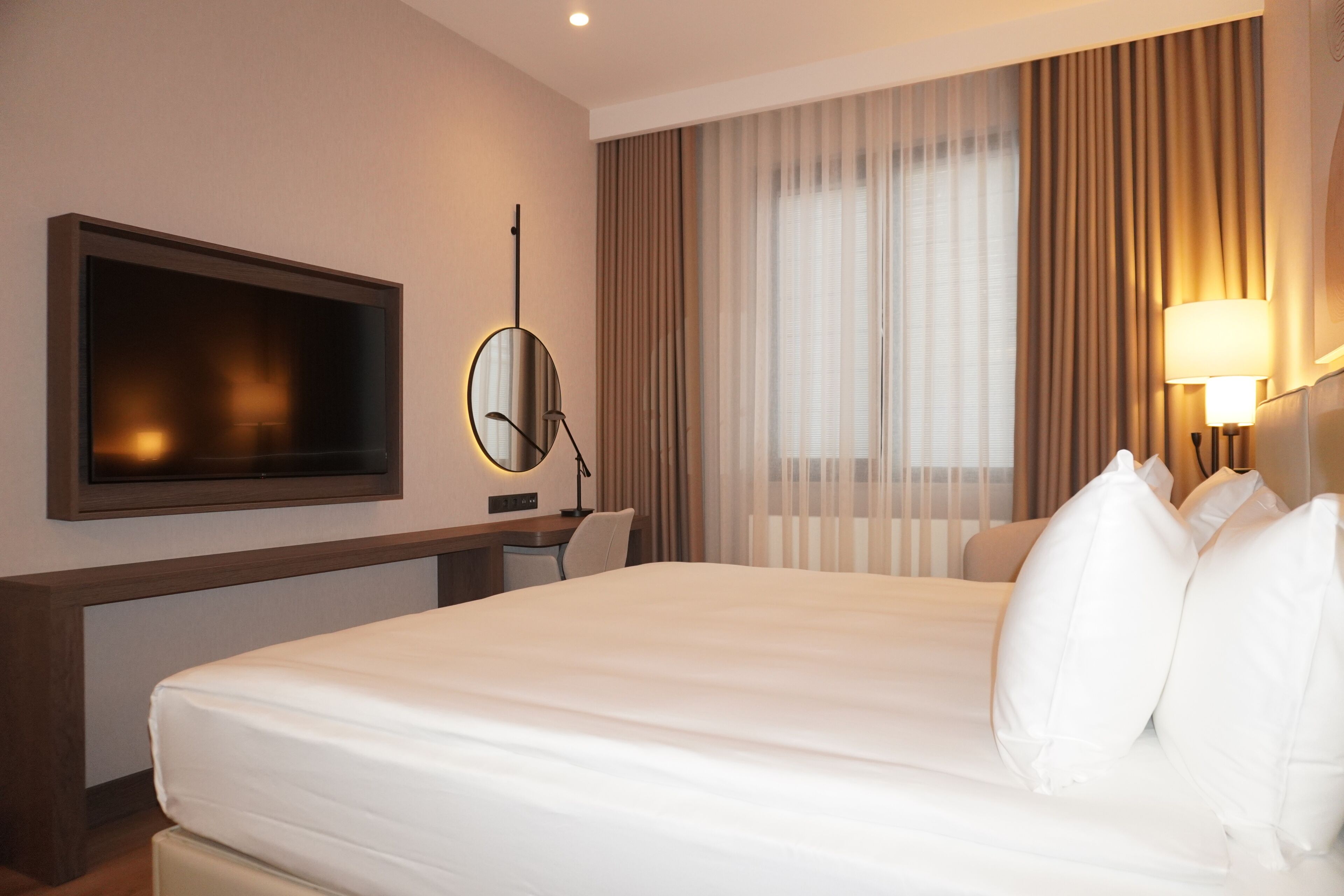 Habitación de hotel con cama, escritorio, televisor, lámpara y ventana con cortinas.