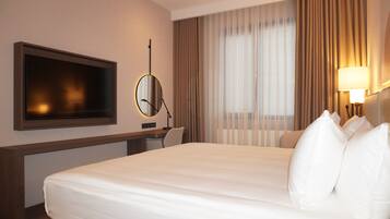 Habitación de hotel con cama, escritorio, televisor, lámpara y ventana con cortinas.