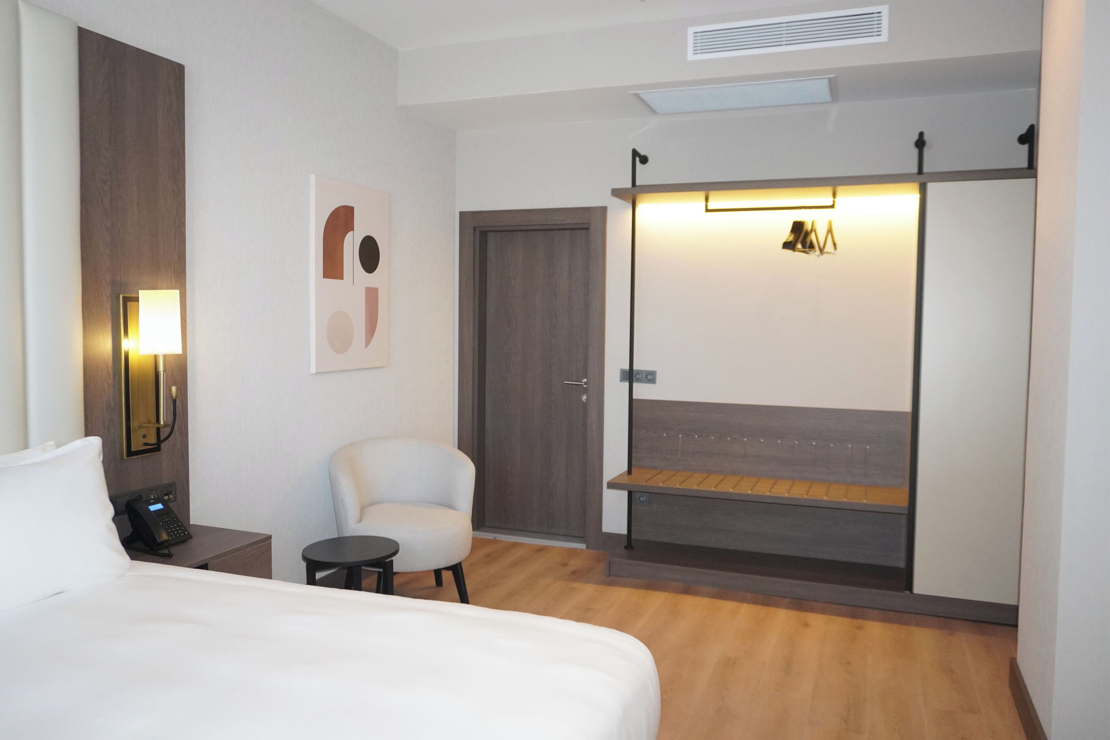 A hotel room with a bed, a chair, a desk with a phone, a coat rack, and a wardrobe.