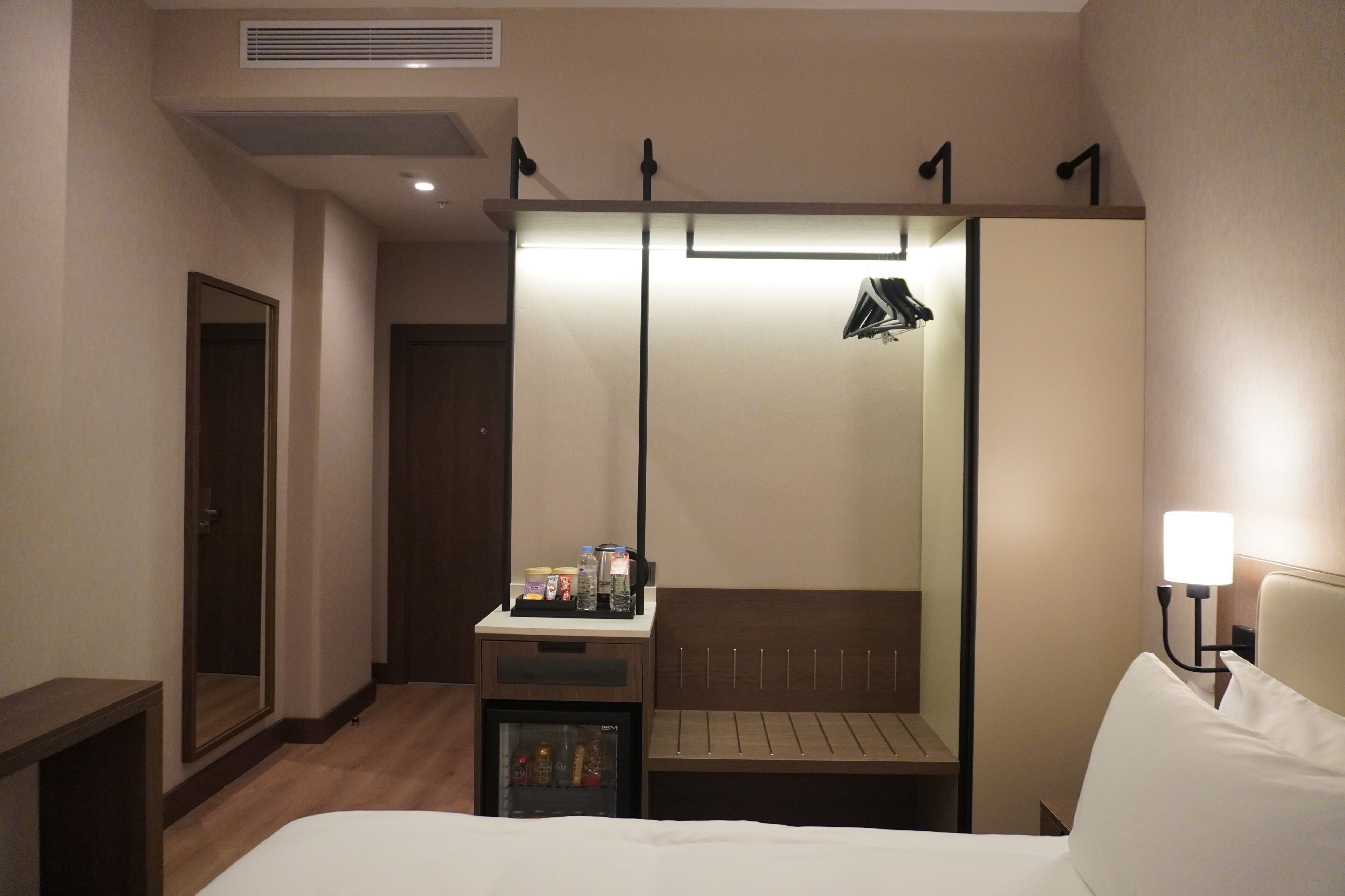 Une chambre d’hôtel équipée d’un lit, d’une table de chevet, d’un miroir, d’une armoire et d’un petit réfrigérateur.