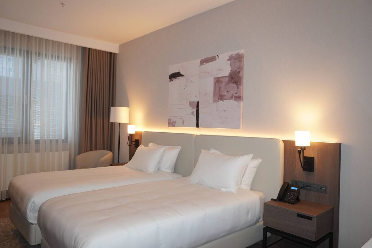 Ein Hotelzimmer mit zwei Betten, einem Schreibtisch, einem Stuhl, einer Lampe, einem Telefon und einem Gemälde an der Wand.