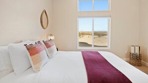 A bedroom with a large bed, two windows, a view of the beach, and a wicker basket with books. - Luxury Desert Retreat on 5 Acres, Hot Tub, BBQ & Stargazing (Joshua Tree)