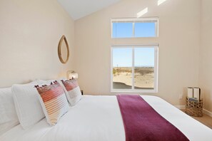Ein Schlafzimmer mit einem großen Bett, zwei Fenstern und Blick auf den Strand, sowie einem Korbwesen mit Büchern.