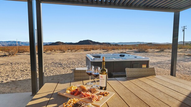 An outdoor patio with a hot tub, a wooden table with food and wine, and a desert landscape in the background. - Luxury Desert Retreat on 5 Acres, Hot Tub, BBQ & Stargazing (Joshua Tree)
