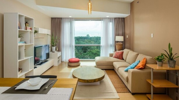 A modern living room with a sofa, a television, a bookshelf, and a large window with a view of greenery. - Modern Luxe Stay: 2BR City Escape (Taguig City)