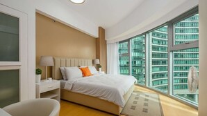 A modern bedroom with a large bed, bedside tables, a desk, and a view of a cityscape through large windows. - Modern Luxe Stay: 2BR City Escape (Taguig City)