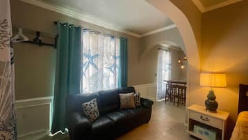 A living room with a black sofa, a small table, a lamp, and a dining area in the background.