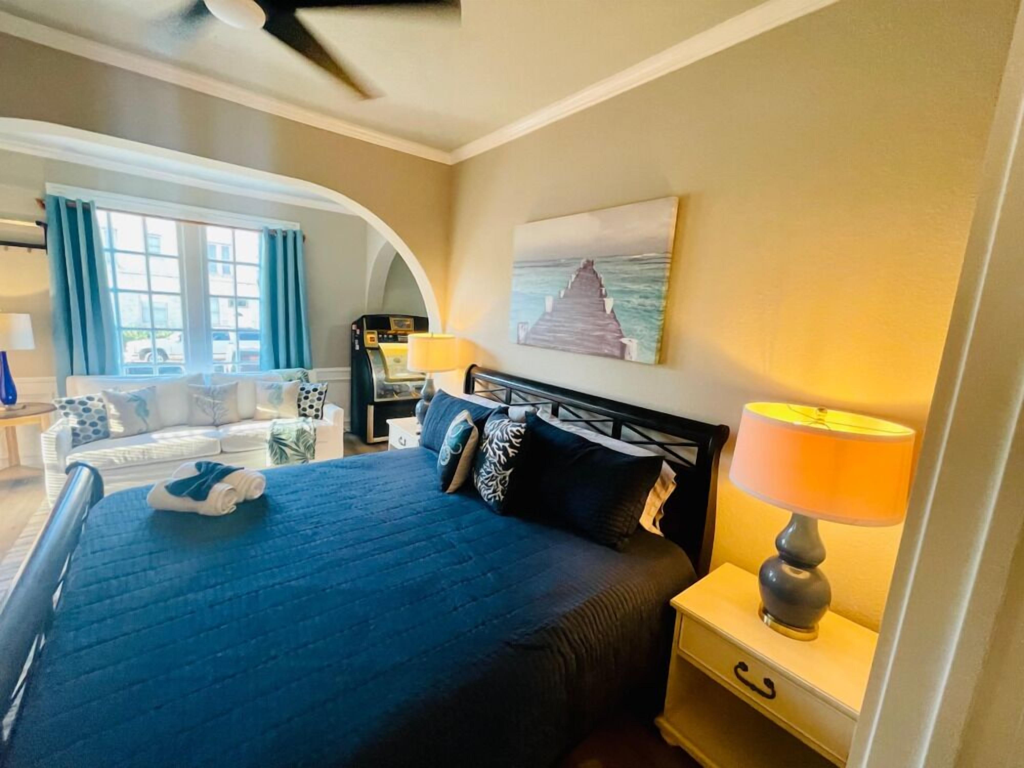 A hotel room with a bed, a nightstand with a lamp, a painting on the wall, and a view of the outdoors through a window.