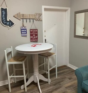 Ein Eckzimmer mit einem weißen Tisch und zwei Stühlen, ein Holzschild mit der Aufschrift 'BEACH!' und 'WORK LIKE A CAPTAIN PLAY LIKE A PIRATE', das an der Wand hängt, sowie ein Spiegel an der Wand.