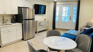 Eine moderne Küche mit weißen Schränken, einem Edelstahlkühlschrank, einem eingebauten Ofen und einem Flachbildfernseher. Es gibt einen Essbereich mit einem runden Tisch und vier Stühlen sowie ein Wohnzimmer mit einem blauen Sofa und gemusterten Kissen.