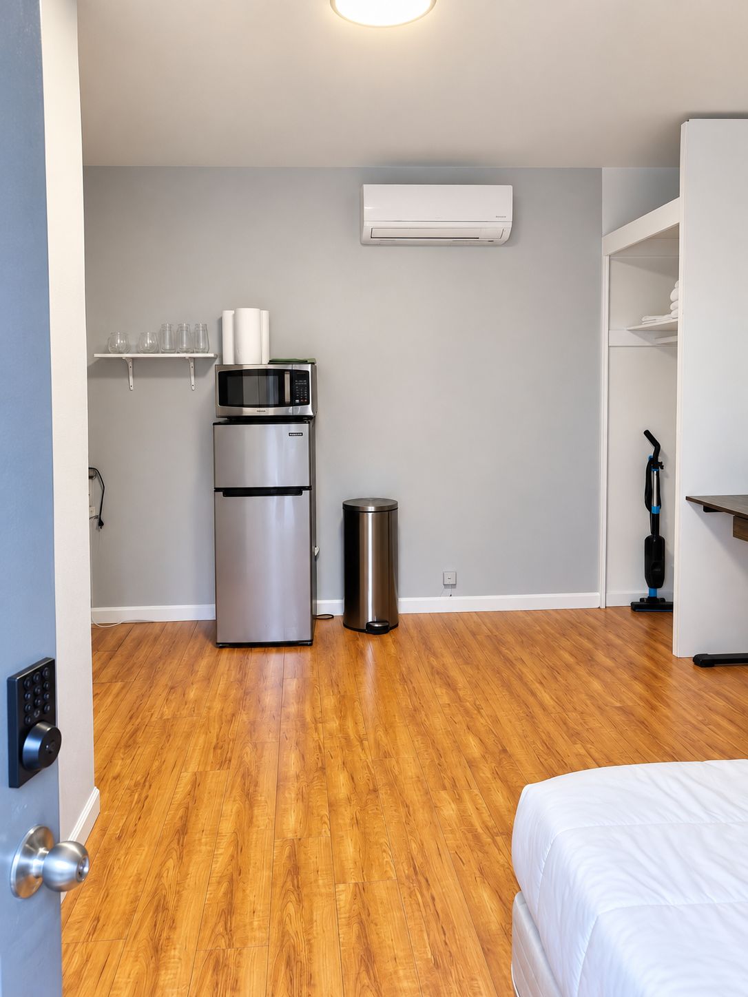 A compact room with a bed, a kitchenette with a refrigerator and microwave, a trash bin, and an air conditioning unit.