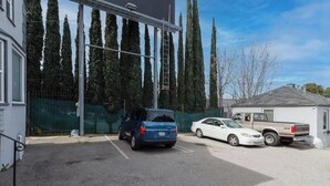 Un estacionamiento con un coche azul y uno blanco, un edificio con valla verde, y árboles altos al fondo.