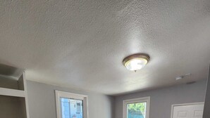Una habitación con suelo de madera, dos ventanas, una puerta y una lámpara en el techo.