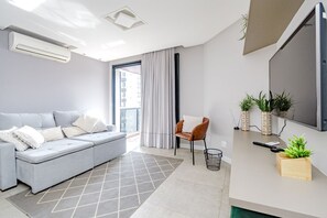 Ein modernes Wohnzimmer mit einem grauen Sofa, einem kleinen Holztisch, einem Fernseher und einem Fenster mit Vorhängen.