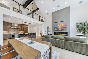 A modern interior with a dining area, kitchen, and living space, featuring a fireplace, large windows, and a mezzanine level. - Stateline Designer Luxury Home (South Lake Tahoe)