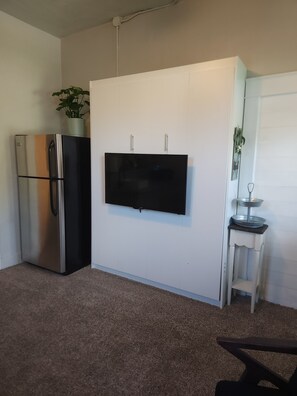 Ein kompakter Wohnraum mit Wandfernseher, Edelstahlkühlschrank und einem kleinen Tisch mit Pflanze.