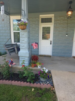 A porch with a white door, a hanging planter with flowers, a flag, and a wind chime.