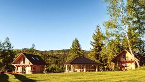 A rustic cabin with a large porch, a smaller cabin with a covered porch, and a wooden structure in a grassy field.