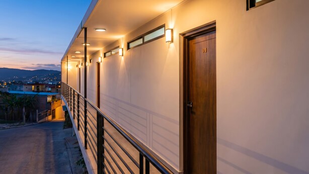 A hotel corridor with a wooden door, a railing, and a view of the city at dusk.