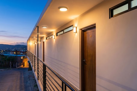 A hotel corridor with a wooden door, a railing, and a view of the city at dusk.