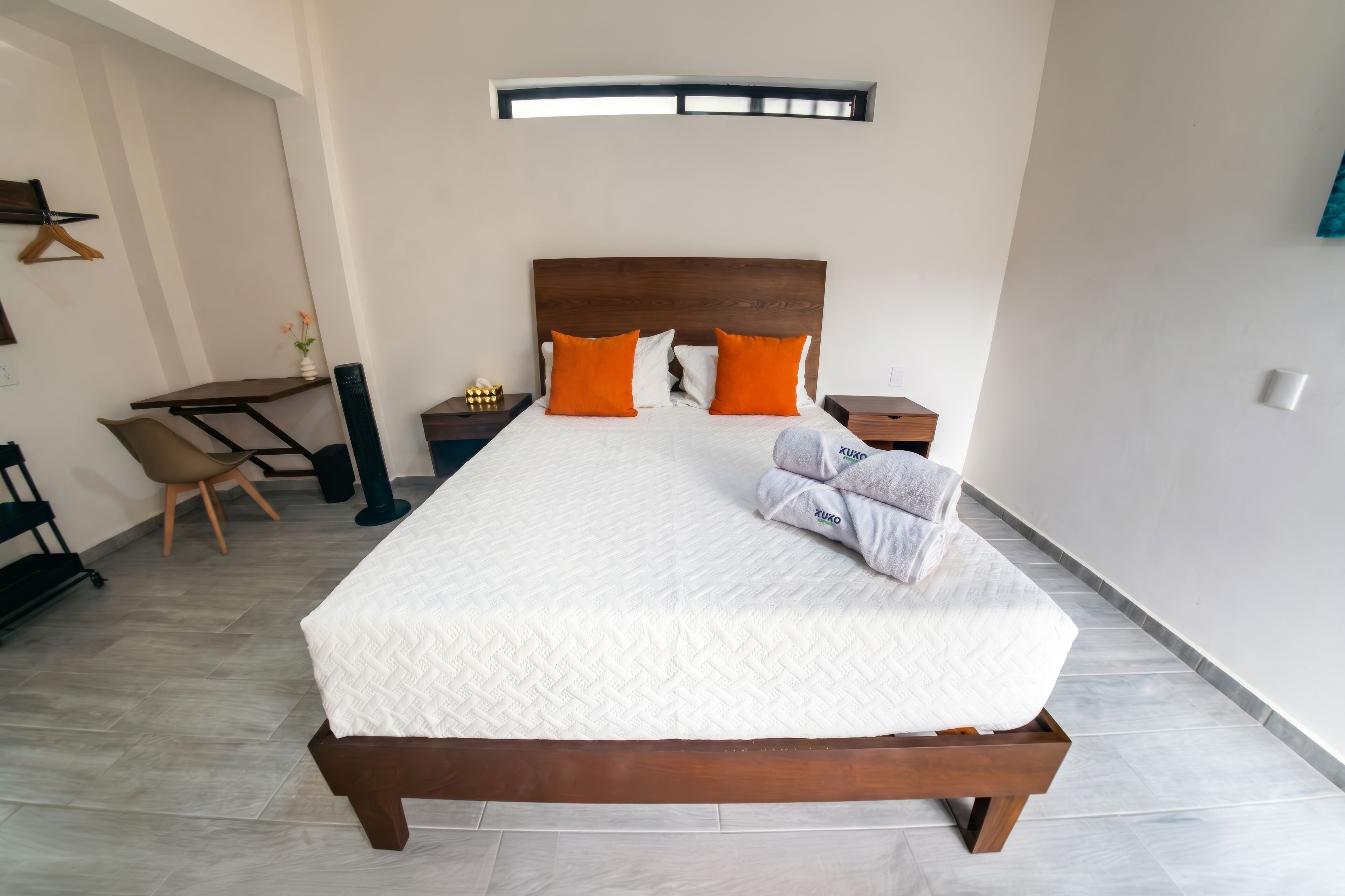 A neatly arranged hotel room with a large bed, a small desk, a chair, and a bedside table with a lamp.