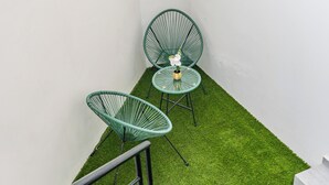 A small outdoor seating area with two green chairs and a table, surrounded by white walls and a patch of artificial grass.