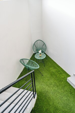 Un pequeño espacio al aire libre con dos sillas verdes y una mesa, rodeado de paredes blancas y una sección de césped artificial.
