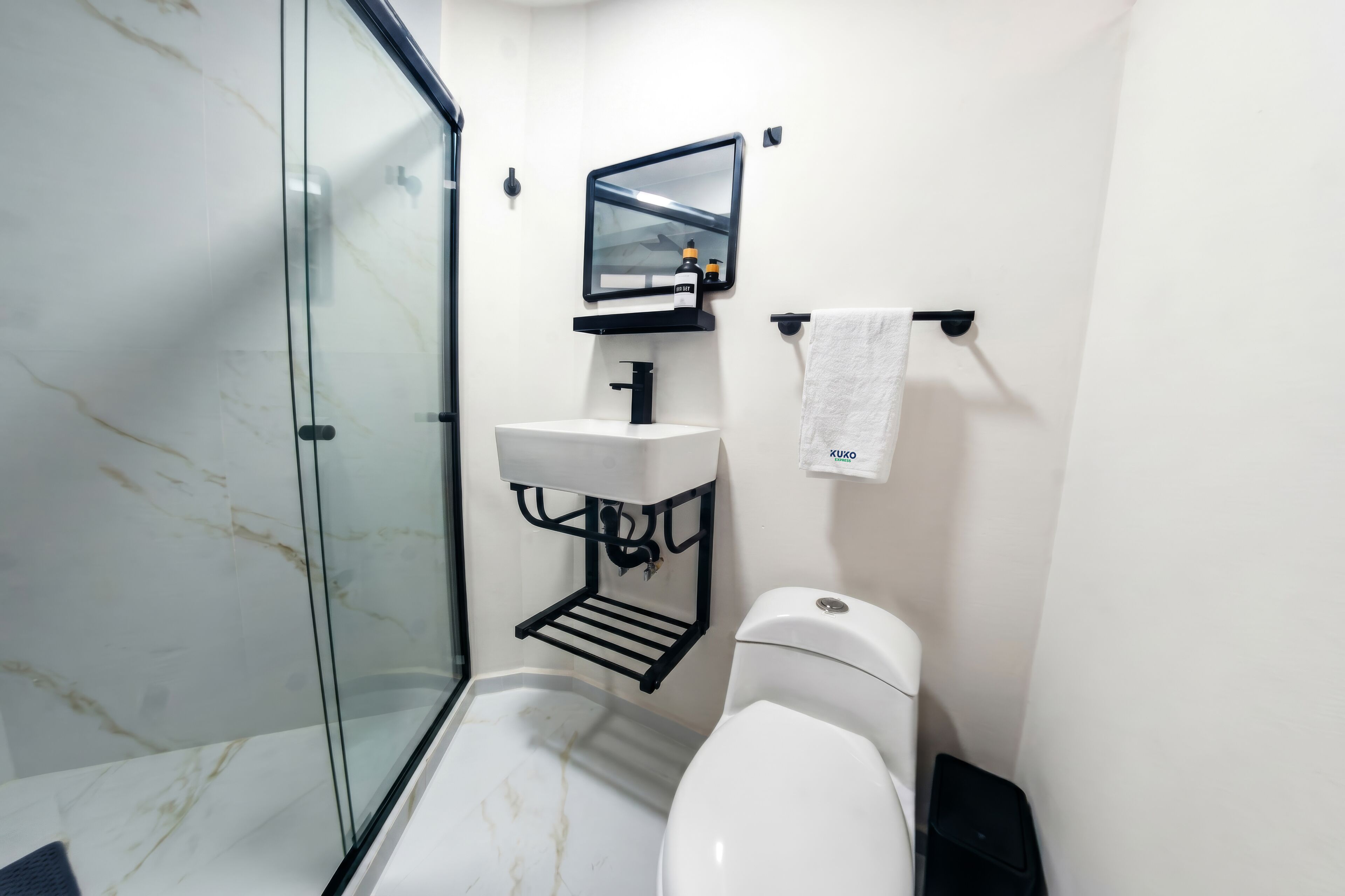 Une salle de bain avec une douche à parois de verre, des toilettes blanches, un lavabo noir avec un miroir au-dessus, et une serviette suspendue à un porte-serviettes.