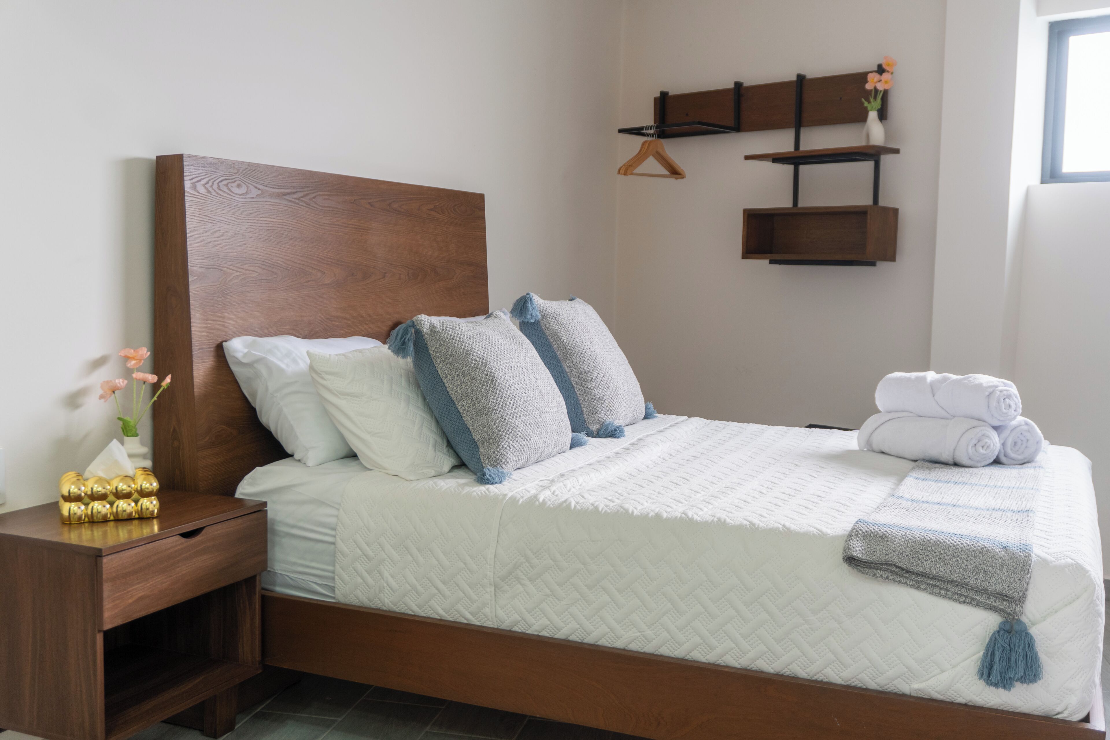 A neatly made bed with a quilted white duvet, two pillows, and a folded towel. A wooden nightstand with a drawer and a vase with flowers is beside the bed.