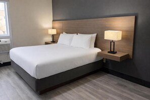 A hotel room with a large bed, two bedside tables with lamps, a dark headboard, and a window with blinds.