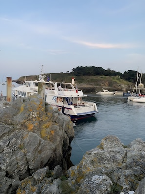 Un port où des bateaux sont amarrés, un paysage rocheux et un phare en arrière-plan.
