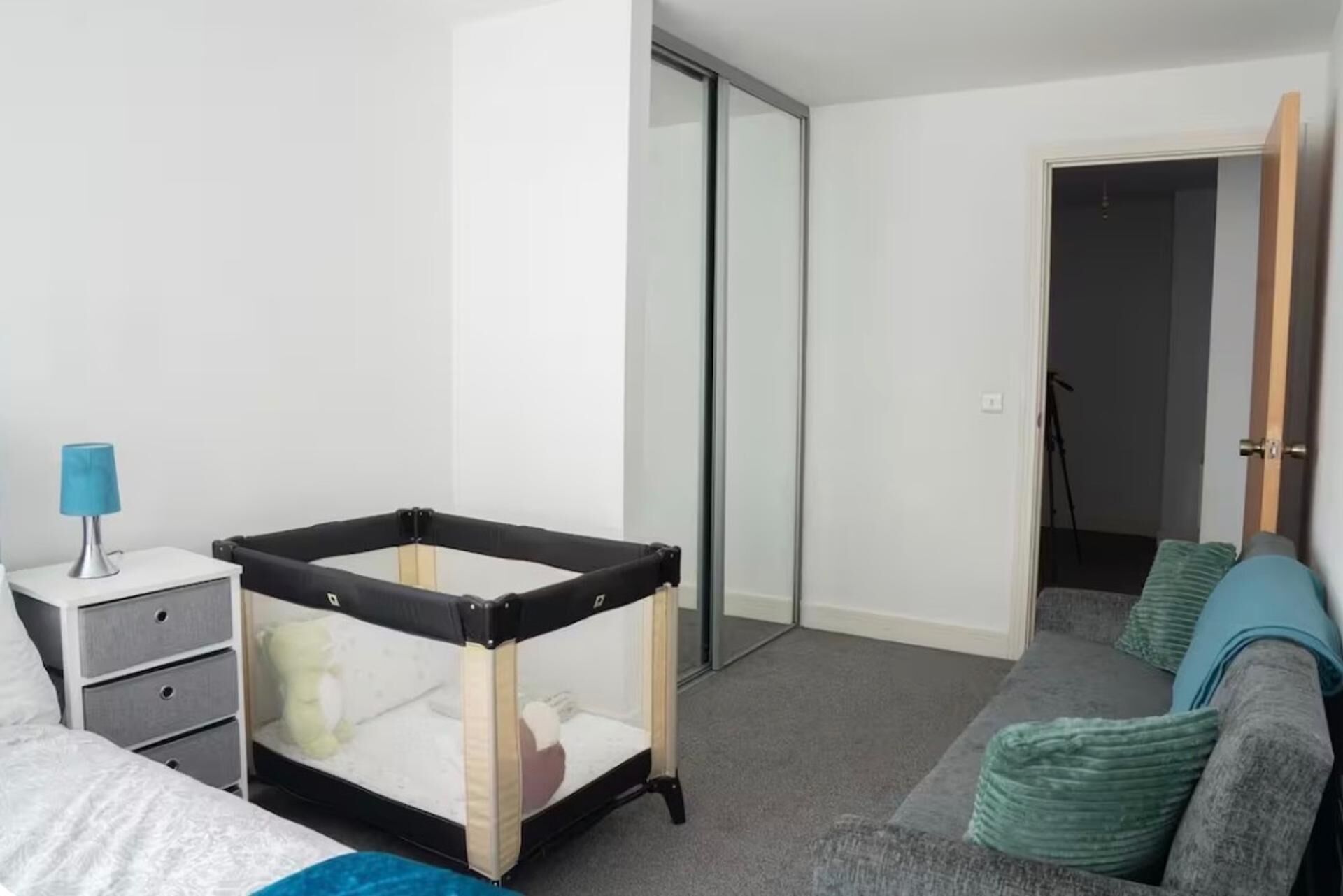 Un dormitorio con cama, una mesita de noche con lámpara, una cuna para bebé blanco y negro, un sofá gris con cojines verdes y una puerta corrediza de vidrio.