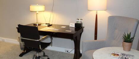 A hotel room with a desk, chair, TV, lamps, and a round table with a magazine.