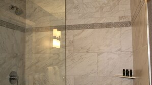 Une salle de bain aux murs recouverts de marbre, dotée d’une douche en verre et d’une étagère où sont rangés les articles de toilette.