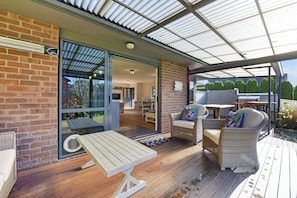 Un espace extérieur abrité doté d’une terrasse en bois, de meubles en osier et d’une porte vitrée donnant accès à un espace intérieur.