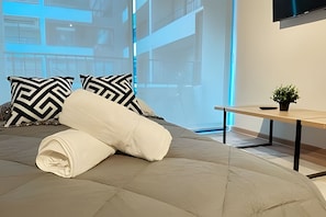 Una cama bien tendida con una toalla blanca enrollada, dos almohadas de diseño blanco y negro, una pequeña planta en maceta sobre una mesita de noche de madera y un amplio ventanal con vista a un edificio.