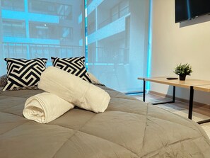 A bed with a rolled white towel, black and white patterned pillows, a wooden nightstand with a small plant, and a TV mounted on the wall. - Moderno depto. en Stgo. cerca del metro (Santiago)