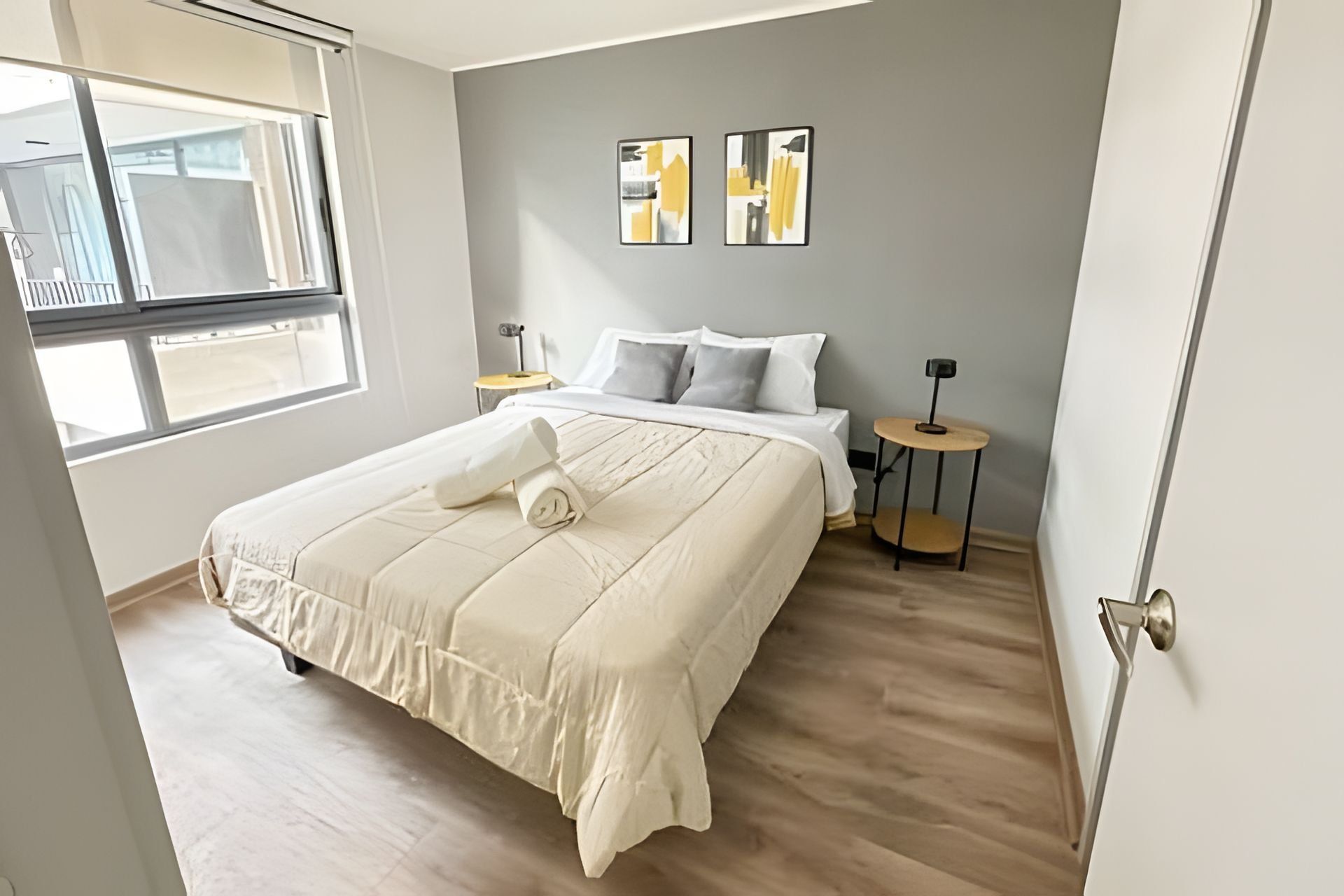 A bedroom with a bed, a small table, a chair, a window, and two wall-mounted picture frames.