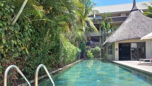 A clear swimming pool with metal handrails, surrounded by lush greenery and a building with a thatched roof. - Pool View Apartment at 'Domaine dès Alizées Club & Spa' (Grand Baie)