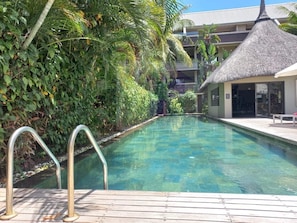 A clear swimming pool with metal handrails, surrounded by lush greenery and a building with a thatched roof. - Pool View Apartment at 'Domaine dès Alizées Club & Spa' (Grand Baie)