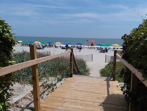 Ein hölzerner Steg führt zu einem Strand mit Sonnenschirmen und Menschen.