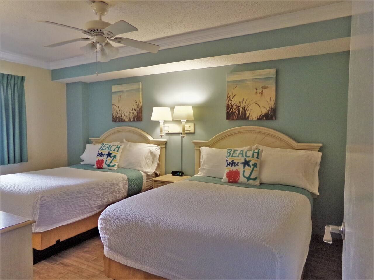 A hotel room with two beds, a ceiling fan, wall art, and a nightstand with a lamp.