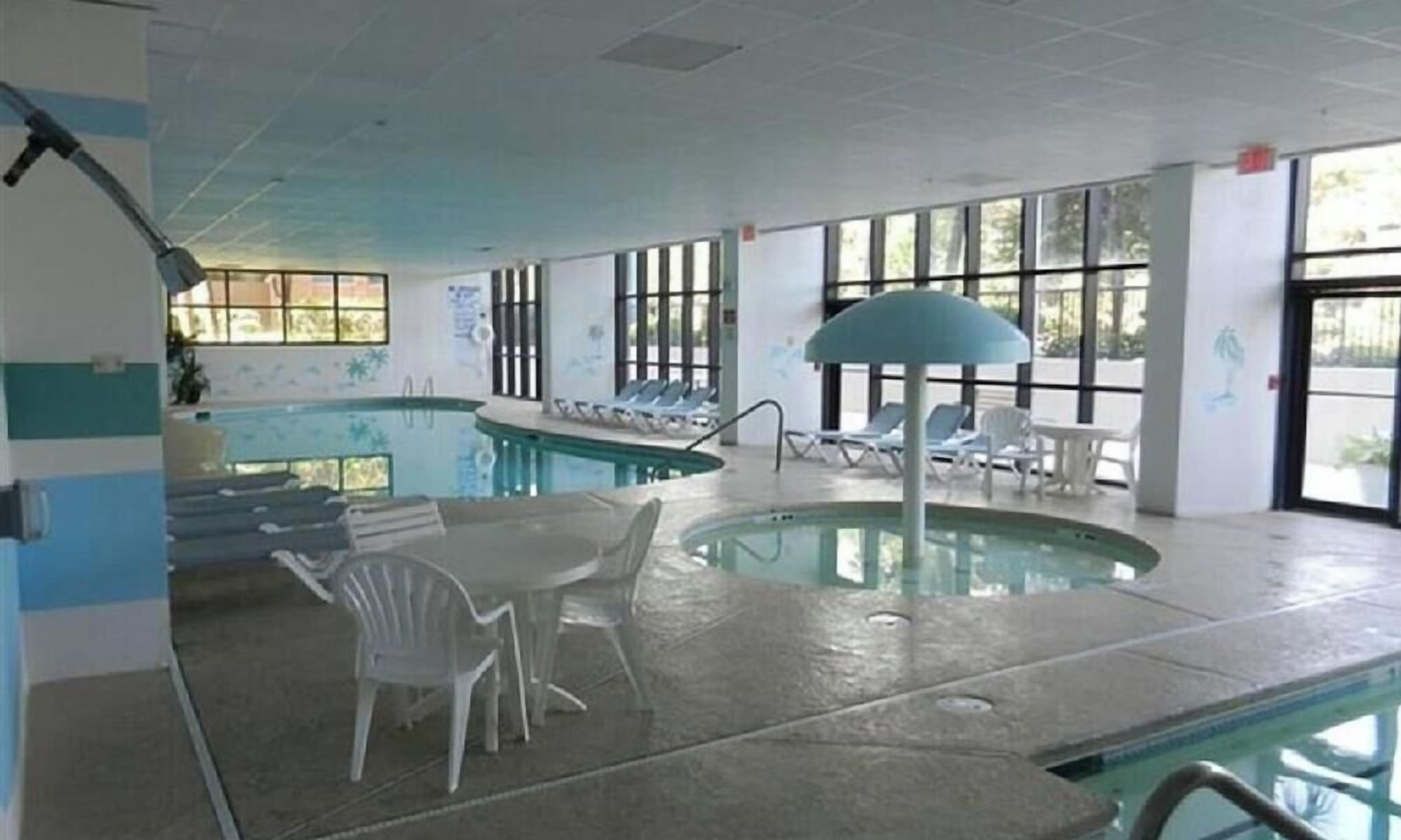 Une piscine intérieure dotée de chaises longues et d’une table, de grandes fenêtres et d’un ventilateur de plafond.