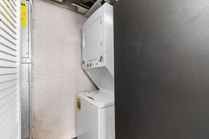 Ein Waschraum mit Waschmaschine und Trockner, einem Lüftungsschacht und einer Tür mit einem gelben Warnschild.