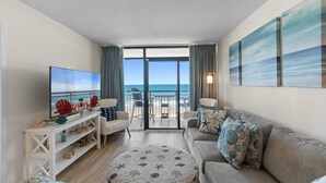 Ein Wohnzimmer mit großem Fernseher, einer Couch, einem runden Hocker und einem Balkon mit Meerblick.
