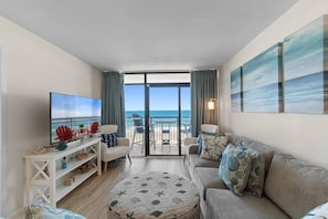 A living room with a large TV, a sofa, a round ottoman, and a balcony with a view of the ocean. - Oceanfront Beach Cove w/ Resort Pools-Wide Balcony (North Myrtle Beach)